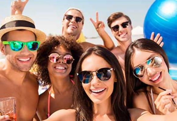 Selfie time! Six cheerful friends in fashionable different swim wear and sun protecting eyewear, caps, with drinks are posing for a selfie photo, that brunette lady is taking, beaming smiles, sunshine